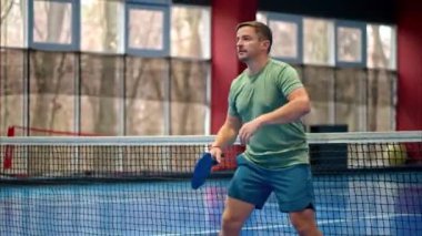 Yeşil tişörtlü bir adam mavi, iç sahada penisbol oynuyor.