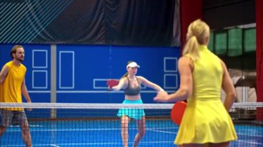 İki erkek ve iki kadın, mavi bir sahada Pickleball oynuyor.