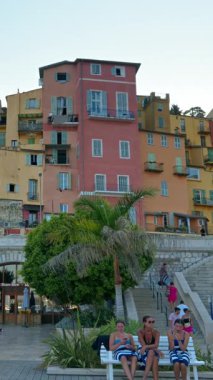 Menton, Fransa - 4 Eylül 2024: Şehirdeki renkli binaların ve kıyı şeridinde yürüyen insanların görüntüsü. Dikey