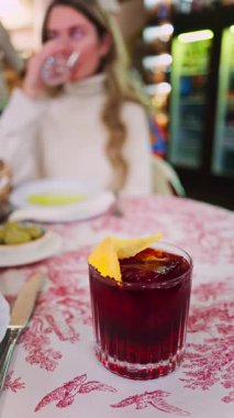 Arka planda bir restoranda içki içen bir kadınla kırmızı ve beyaz masa örtüsünün üzerinde negroni kokteyli yakın plan. Dikey