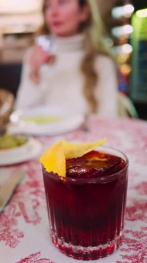 Arka planda bir restoranda içki içen bir kadınla kırmızı ve beyaz masa örtüsünün üzerinde negroni kokteyli yakın plan. Dikey