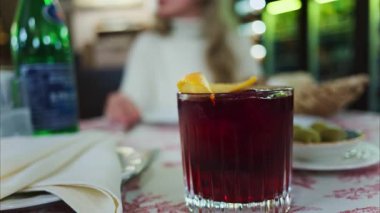 Arka planda bir restoranda oturan bir kadınla kırmızı ve beyaz bir masa örtüsünün üzerine negroni kokteylini yakın plan. Dikey