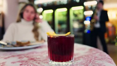 Kırmızı ve beyaz bir masa örtüsü üzerine negroni kokteyli yakın plan arkasında bir restoranda oturan bir kadın