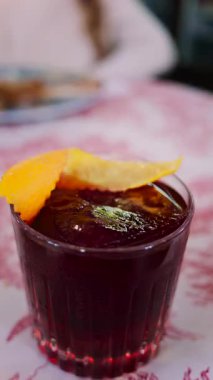 Arka planda bir restoranda içki içen bir kadınla kırmızı ve beyaz masa örtüsünün üzerinde negroni kokteyli yakın plan. Dikey