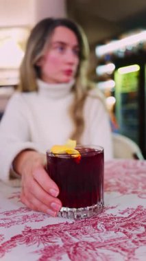 Lokantada kırmızı beyaz masa örtüsüne negroni kokteyli koyan bir kadının yakın plan fotoğrafı. Dikey