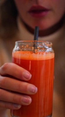 Bir kadının portakal ve havuç suyunu tahta bir masada siyah bir pipetle içerken yakın plan fotoğrafı. Dikey