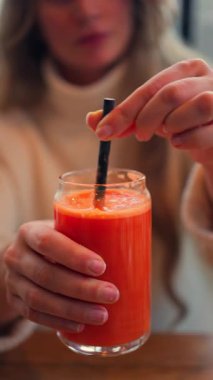 Bir kadının portakal ve havuç suyunu bir bardakla ahşap bir masada siyah bir pipetle karıştırması. Dikey