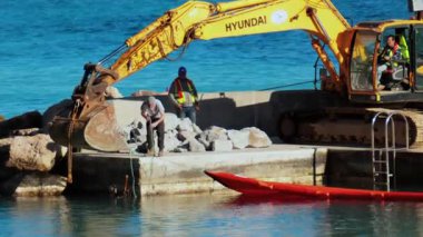 Nice, Fransa - 10 Şubat 2025: Büyük, sarı bir kazıcı ve arka planda deniz olan kırmızı yüzen bir bariyerle ilgilenen bir işçi