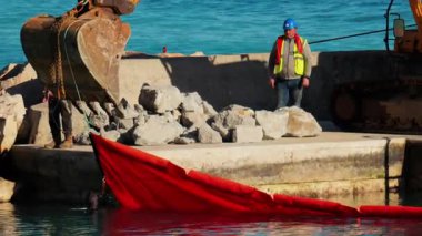 Nice, Fransa - 10 Şubat 2025: Büyük, sarı bir kazıcı ve arka planda deniz olan kırmızı yüzen bir bariyerle ilgilenen bir işçi