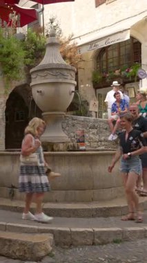 Saint Paul de Vence, Fransa - 10 Ağustos 2024: İnsanlar şehrin caddesinde bir çeşmenin yanında yürüyorlar. Dikey