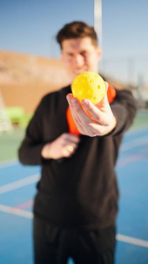 Genç, mutlu ve gülümseyen bir adamın portatifi. Pickleball raketi ve kortta bir top tutuyor.