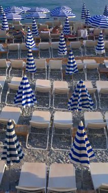 Nice, Fransa - 18 Ağustos 2024: İnsanlar mavi ve beyaz çizgili şemsiyeler altında sahilde yüzüyor ve dinleniyorlar. Dikey