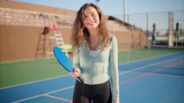 Kıvırcık saçlı, mutlu ve gülümseyen bir kadın, elinde bir Pickleball mavi raketi ve güneşli bir günde mavi kortta sarı bir top tutuyor.