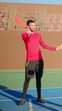 Kırmızı tişörtlü bir adam güneşli bir günde mavi kortta nasıl top oynanacağını öğretiyor. Dikey
