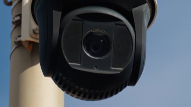 Yuvarlak, siyah güvenlik kamerası beyaz bir direğe monte edilmiş ve arka planda gökyüzü var.