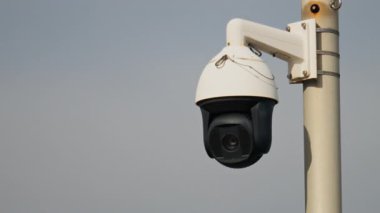 Yuvarlak, siyah beyaz bir güvenlik kamerasının yakın plan üzerinde bulutlu bir gökyüzü olan beyaz bir direğe monte edilmesi.