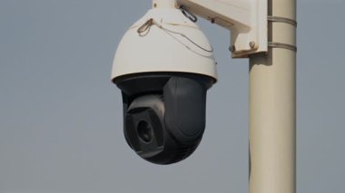 Yuvarlak, siyah beyaz bir güvenlik kamerasının yakın plan üzerinde bulutlu bir gökyüzü olan beyaz bir direğe monte edilmesi.