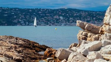 Kıyıdaki kayalıklar Juan-les-Pins 'de denizde ilerleyen bir yelkenli yelkenli, Fransa' da şehir ve arka planda dağ.