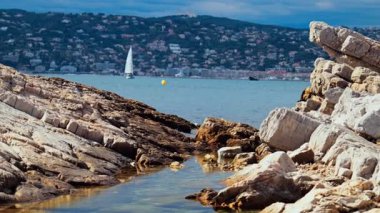 Kıyıdaki kayalıklar Juan-les-Pins 'de denizde ilerleyen bir yelkenli yelkenli, Fransa' da şehir ve arka planda dağ.