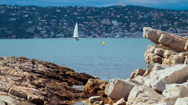 Kıyıdaki kayalıklar Juan-les-Pins 'de denizde ilerleyen bir yelkenli yelkenli, Fransa' da şehir ve arka planda dağ.