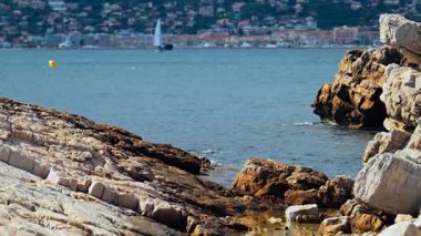 Kıyıdaki kayalıklar Juan-les-Pins 'de denizde ilerleyen bir yelkenli yelkenli, Fransa' da şehir ve arka planda dağ.