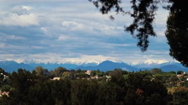 Yeşil ağaçlarla çevrili turuncu villalar ve bulutlu bir günde arka planda dağlar.