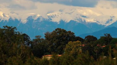 Yeşil ağaçlarla çevrili turuncu villalar ve bulutlu bir günde arka planda dağlar.