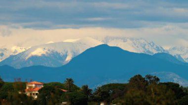 Yeşil ağaçlarla çevrili turuncu villalar ve bulutlu bir günde arka planda dağlar.