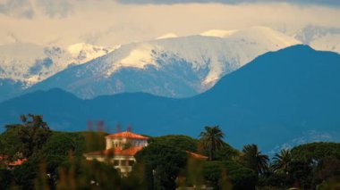 Yeşil ağaçlarla çevrili turuncu villalar ve bulutlu bir günde arka planda dağlar.