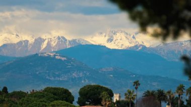 Yeşil ağaçlarla çevrili turuncu villalar ve bulutlu bir günde arka planda dağlar.