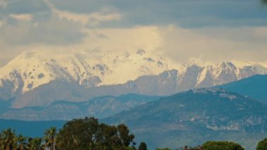 Yeşil ağaçlarla çevrili turuncu villalar ve bulutlu bir günde arka planda dağlar.
