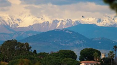 Bulutlu bir günde yeşil ağaçlarla çevrili binaların üzerinde uçan bir uçağın uzak görüntüsü.