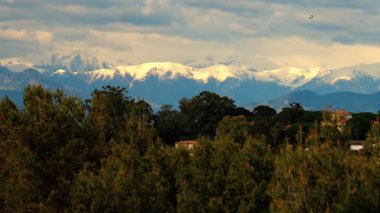 Yeşil ağaçlarla çevrili turuncu villalar ve bulutlu bir günde arka planda dağlar.