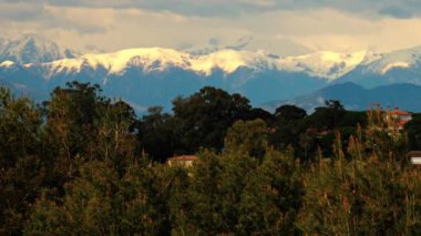 Yeşil ağaçlarla çevrili turuncu villalar ve bulutlu bir günde arka planda dağlar.