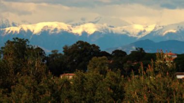 Yeşil ağaçlarla çevrili turuncu villalar ve bulutlu bir günde arka planda dağlar.