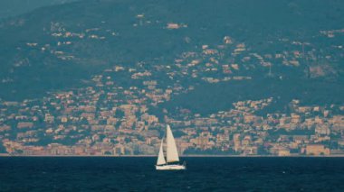 Arka planda binalar ve dağlarla denizde hareket eden bir yelkenlinin uzak görüntüsü
