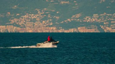 Hareketli bir teknede insanlar binalar ve arkası dağlarla dolu.