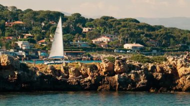 Denizde ilerleyen beyaz bir yelkenli görüntüsü, kayaların yanında ve arka planda kasaba.