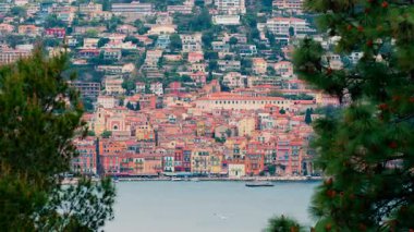 Close up of green tree branches with a blurred, distant view of the colourful buildings of Villefranche-sur-Mer, France