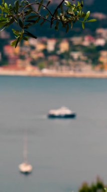 Close up of a green tree branch with a blurred view of the colourful buildings Villefranche-sur-Mer, France and a boat moving on the sea. Vertical