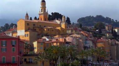 Menton, Fransa St Michel Bazilikası 'nın uzak manzarası bulutlu bir günde renkli binalar ve palmiye ağaçları ile çevrilidir.