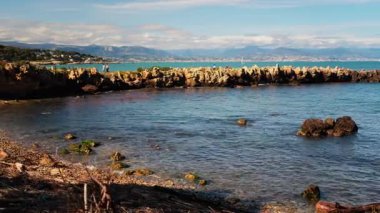 Dalgalar kayalara çarpıyor ve insanlar arka planda yürüyor.