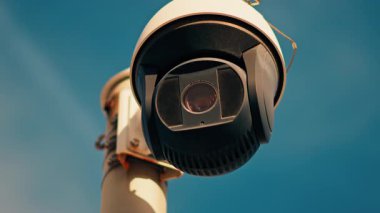 Yuvarlak, siyah beyaz bir güvenlik kamerasının yakın plan üzerinde gökyüzü olan beyaz bir direğe monte edilmiş.