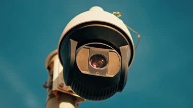 Yuvarlak, siyah beyaz bir güvenlik kamerasının yakın plan üzerinde gökyüzü olan beyaz bir direğe monte edilmiş.