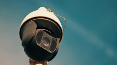 Yuvarlak, siyah beyaz bir güvenlik kamerasının yakın plan üzerinde gökyüzü olan beyaz bir direğe monte edilmiş.