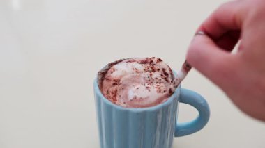 Mavi bir fincan sıcak çikolatayı beyaz bir masada karıştıran bir adamın elini kapat.