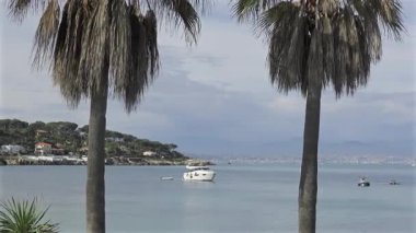 Mavi denizde kayıkları olan palmiye ağaçları ve kıyı kasabası ve arka planda dağlar.