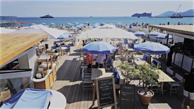 Cannes, Fransa - 28 Nisan 2025: İnsanlar bir sahil restoranında güneş şemsiyesi altında yürüyorlar