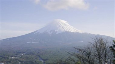 Fuji Dağı açık mavi gökyüzüne karşı karla kaplı bir zirve ile dimdik ayakta duruyor. Çevresindeki manzarada canlı bahar yaprakları sergileniyor.