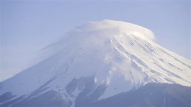 A breathtaking sight of a snow-covered volcano as clouds gracefully swirl around its peak during the sunset light, showcasing nature's beauty in serene surroundings.